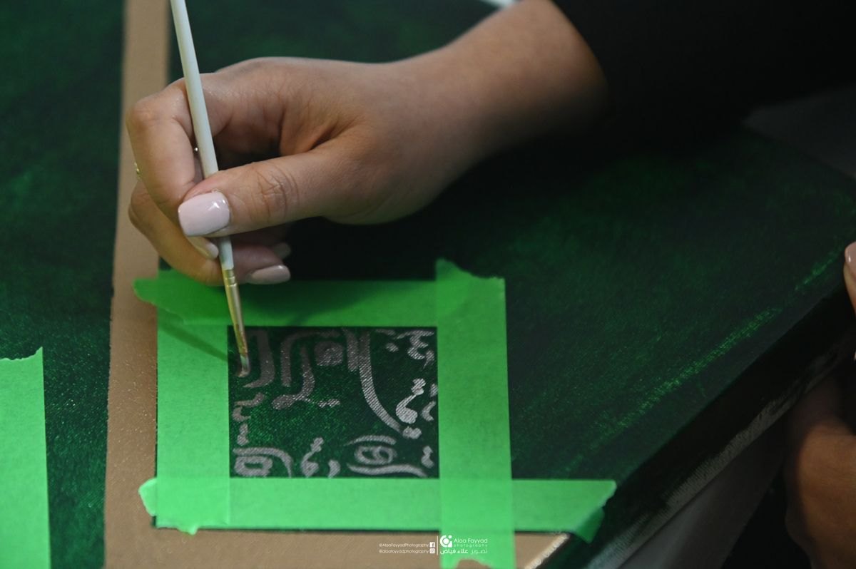 Process detail — Hala Ali applying calligraphy with masking tape on green canvas